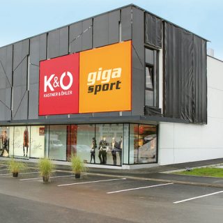 Modernes Gebäude mit großen Schildern für K&Ö Kastner & Öhler und Giga Sport. Das Gebäude ist minimalistisch gestaltet und kombiniert Glas und Paneele. In den Fenstern sind Schaufensterpuppen in Sportbekleidung zu sehen. Im Vordergrund ist ein Parkplatz mit einigen Zierpflanzen zu sehen.