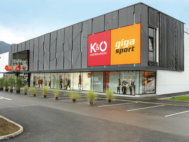 Un moderno edificio commerciale con un grande cartello rosso-arancione con la scritta "K&O Kastner & Öhler" e "giga sport". La facciata esterna è dotata di pannelli scuri e grandi vetrine che espongono manichini. In primo piano si vede un piccolo parcheggio.
