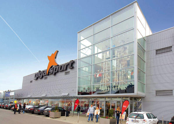 Außenansicht eines großen Geschäfts mit Glasfassade. Das Gebäude trägt in fetten Buchstaben das Logo und den Namen „Gigasport“. Davor parken mehrere Autos, und unter einem klaren blauen Himmel gehen Menschen durch den Haupteingang ein und aus.