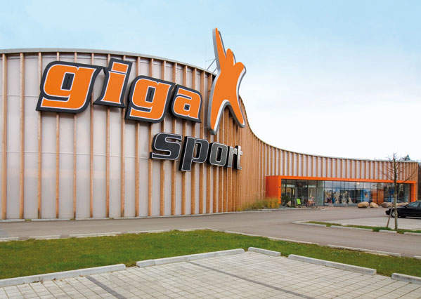 Ein großes Gebäude mit „gigaX sport“ in fetten orangefarbenen und schwarzen Buchstaben auf der Fassade. Das Gebäude hat ein modernes Design mit einem geschwungenen Dach. Im Vordergrund sind ein Parkplatz und ein kleiner Rasenfleck zu sehen.