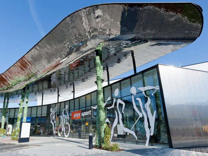 Modernes Gebäudeäußeres mit großem, reflektierendem Überhang und Glasfassade. Auf dem Glas sind weiße Silhouetten von Sportlern in Bewegung zu sehen. Verschiedene Pflanzen schmücken den Bereich und auf einem Schild steht in Rot und Weiß „gigasport“.