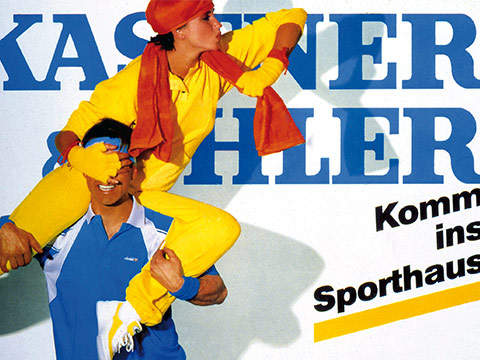 Ein Mann im blau-weißen Sporthemd trägt eine Person, die in leuchtendem Gelb und Rot gekleidet ist. Im Hintergrund ist ein großer, teilweise sichtbarer blauer Text zu sehen, in kleinerem schwarzen Text steht „Komm ins Sporthaus“.