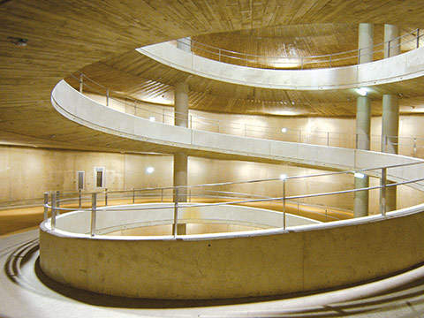 Ein moderner, spiralförmiger Betoninnenraum mit einem gewundenen Weg und Geländern. Die Wände und Decken bestehen aus glattem Beton und der Raum wird mit sanftem, warmem Licht beleuchtet.