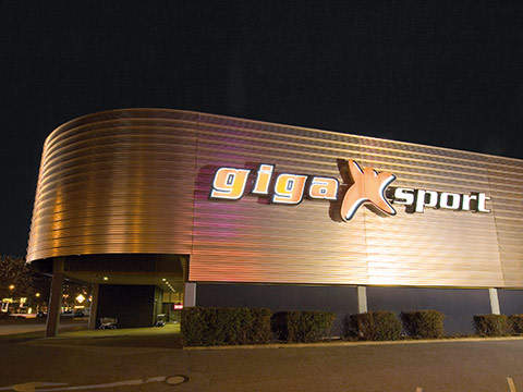 Ein modernes Gebäude mit geschwungener Fassade und einem großen beleuchteten Schild mit der Aufschrift „gigaXsport“ vor einem dunklen Himmel. Das Äußere ist metallisch und warm beleuchtet. Büsche säumen die Vorderseite des Gebäudes.