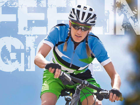 Ein Radfahrer in blau-grüner Kleidung mit Helm und Sonnenbrille fährt Fahrrad. Im Hintergrund ist ein großer, teilweise sichtbarer Text mit einem blauen Himmel dahinter zu sehen.