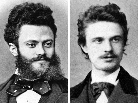 Zwei Vintage-Porträts in Sepiatönen von Männern in formeller Kleidung. Der Mann links hat einen Vollbart und lockiges Haar, während der Mann rechts einen Schnurrbart und welliges Haar hat. Beide tragen dunkle Anzüge mit Fliege.