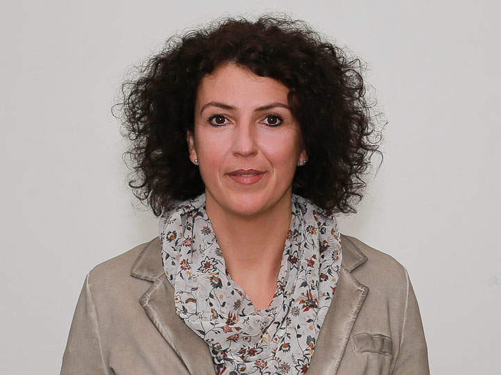 Una persona con capelli scuri e ricci, che indossa una sciarpa a fiori e un blazer beige, guarda direttamente la fotocamera. Lo sfondo è semplice e neutro.