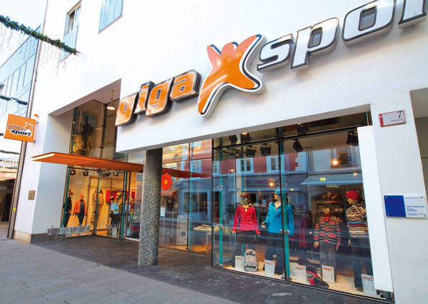 Schaufenster eines Sportartikelgeschäfts mit großen Glasfenstern, in denen Schaufensterpuppen in Sportbekleidung zu sehen sind. Der Name „Gigasport“ prangt in großen Lettern prominent über dem Eingang.