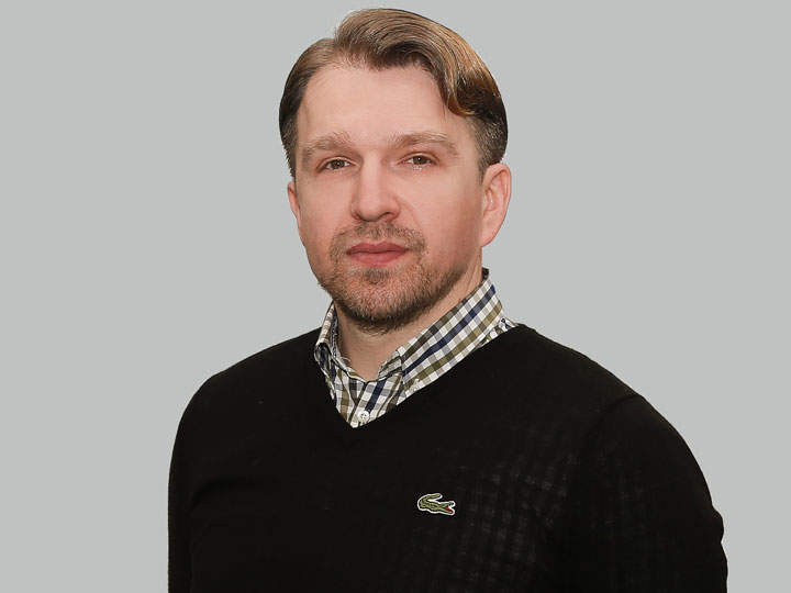 Un uomo con capelli corti e barba indossa un maglione nero sopra una camicia a quadri. È in piedi davanti a uno sfondo grigio semplice e guarda la fotocamera.