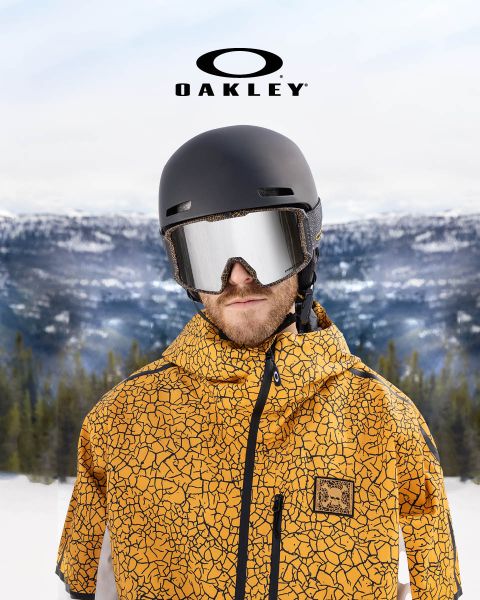 Una persona con casco nero, occhiali protettivi riflettenti e giacca gialla con motivo a increspatura si trova di fronte a un paesaggio montano innevato. Nella parte superiore dell'immagine si vede il logo Oakley.