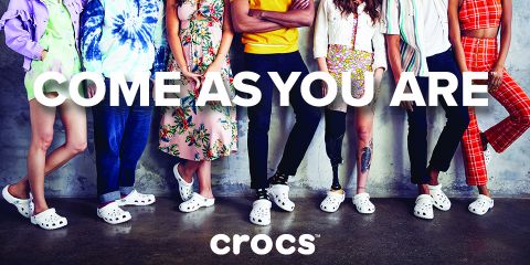 Un gruppo eterogeneo di persone è in piedi accanto a un muro di cemento. Ognuno indossa un outfit diverso e Crocs bianche. Il testo "COME AS YOU ARE" è ben visibile sopra il logo Crocs.