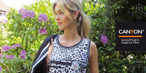 Una donna in un top senza maniche con un motivo bianco e nero è in piedi all'aperto accanto a fiori viola in fiore e piante verdi. Sul lato destro si vede il logo "CANYON women sports". Tiene una giacca nera sulla spalla.