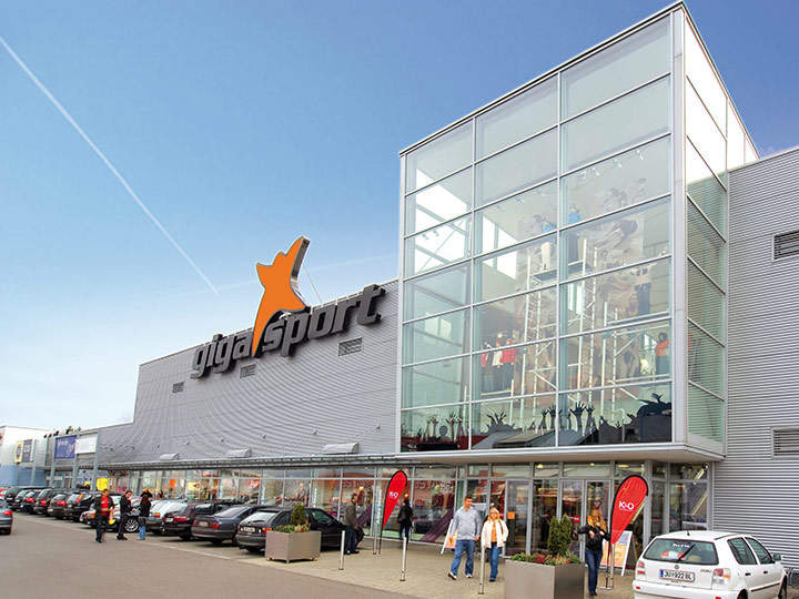 Vista esterna di un grande negozio di articoli sportivi con facciata in vetro e logo arancione. Diverse auto sono parcheggiate nel parcheggio e alcune persone si dirigono verso l'ingresso. L'edificio ha un design moderno sotto un cielo azzurro limpido.