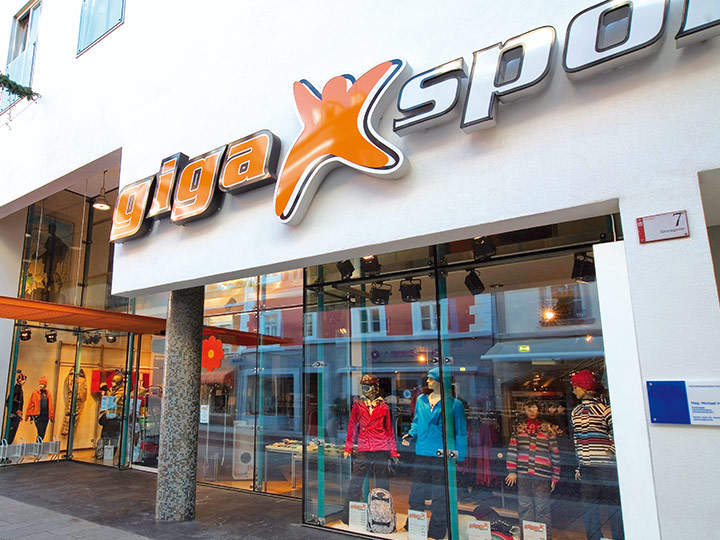 Vista esterna di un negozio Gigasport con una facciata moderna. L'ingresso è fiancheggiato da ampie vetrate che espongono manichini vestiti con abbigliamento sportivo colorato. Sopra la porta è affissa un'insegna ben visibile con la scritta "Gigasport".