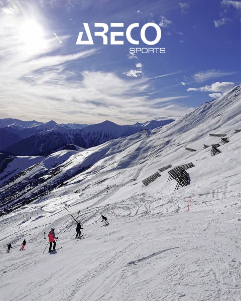 Sciatori su un pendio montuoso innevato sotto un cielo blu con nuvole sparse. Il sole splende luminoso e proietta ombre sulla neve. In alto si legge il testo "ARECO SPORTS". Sullo sfondo si vedono cime innevate.
