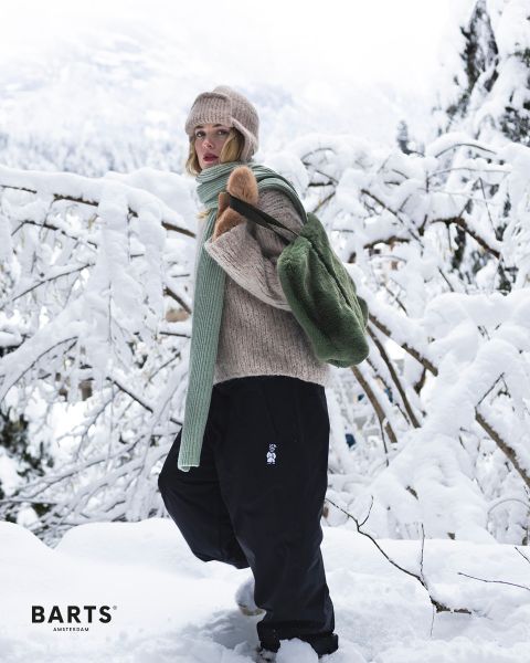Una persona con un berretto di maglia beige, un maglione, una sciarpa verde e pantaloni neri si trova in un paesaggio innevato e indossa uno zaino verde. Rami e alberi innevati caratterizzano lo sfondo. Il testo sull'immagine è "BARTS AMSTERDAM".