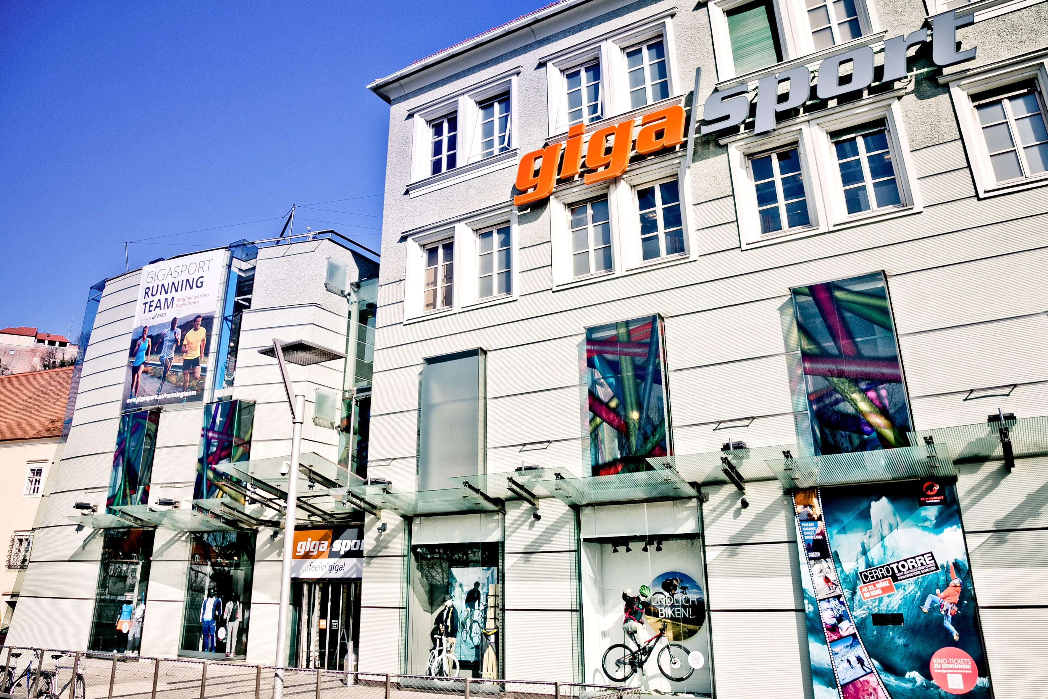 Außenansicht eines Gigasport-Geschäfts mit großen Fenstern, auf denen Sportbilder zu sehen sind. Das Gebäude ist mit einem auffälligen orangefarbenen „Gigasport“-Schild versehen. Ein Glasdach schützt den Eingang vor hellem Tageslicht.