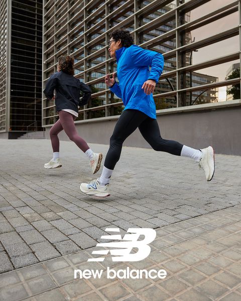 Due persone fanno jogging all'aperto, una con una giacca blu e leggings neri, l'altra con una giacca nera e leggings color castagna. Entrambe indossano scarpe da ginnastica bianche. Corrono accanto a un edificio moderno con pannelli di vetro. In basso si vede il logo New Balance.