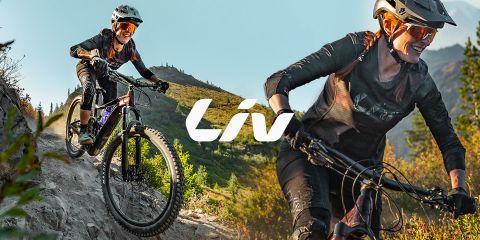 Due donne vanno in mountain bike su un sentiero accidentato con alberi e montagne sullo sfondo. Entrambe indossano caschi e occhiali da sole. Al centro si vede chiaramente la parola "Liv".