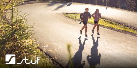 Due persone fanno jogging su una strada tortuosa circondata da una foresta. Il sole proietta lunghe ombre sul marciapiede. Entrambi indossano abbigliamento sportivo. Nell'angolo in basso a sinistra c'è un logo con la scritta "Stuf".