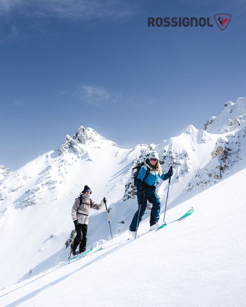 Due sciatori in abbigliamento invernale salgono su un pendio montuoso innevato in una giornata limpida. Sullo sfondo si vedono cime frastagliate e innevate contro un cielo blu. Il logo Rossignol è visibile nell'angolo in alto a destra.
