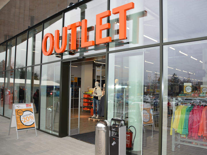 Schaufenster mit großem „OUTLET“-Schild über dem Eingang. Im Inneren sind Schaufensterpuppen und Kleiderständer zu sehen. Ein Schild draußen wirbt für 20 % Rabatt. Glasfenster zeigen Spiegelungen und einen Blick auf den Parkplatz.