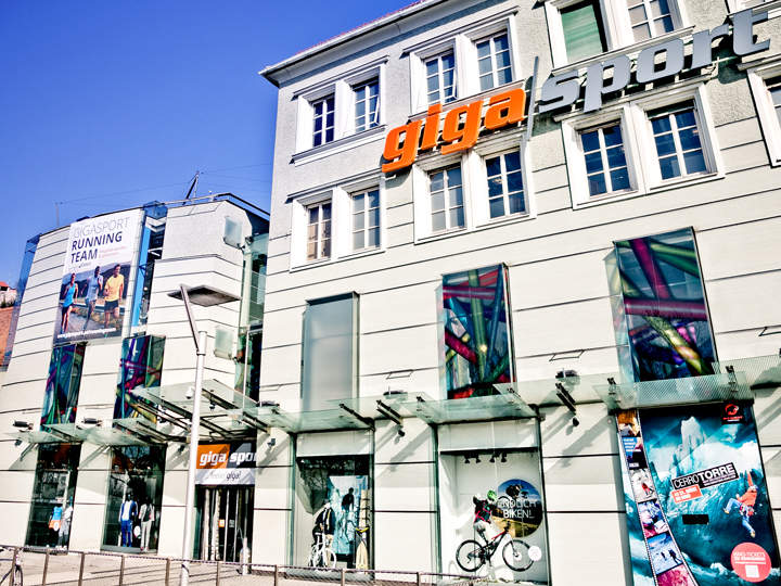 Vista esterna del negozio Gigasport con ampie vetrate che espongono articoli sportivi. Un cartellone pubblicitario mostra dei corridori. L'edificio ha una facciata moderna con la scritta "Gigasport" in audaci lettere arancioni e grigie.