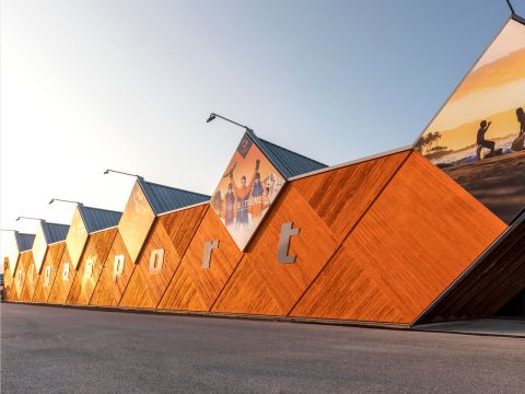 Edificio moderno con facciata esagonale in legno e la scritta "ggysport". Nella parte superiore si notano grandi poster, tra cui immagini di persone che praticano attività all'aperto. Sullo sfondo si vede un cielo azzurro brillante.