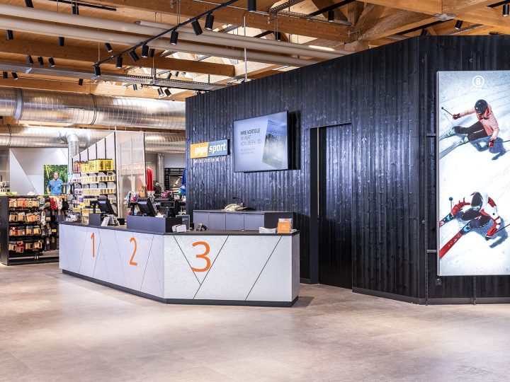 Un interno moderno di un negozio di articoli sportivi con un elegante banco di assistenza clienti con le scritte 1, 2, 3. Sulla parete dietro si vedono immagini sportive e ci sono scaffali ben ordinati con vari articoli sportivi. Il soffitto presenta travi e tubature a vista.