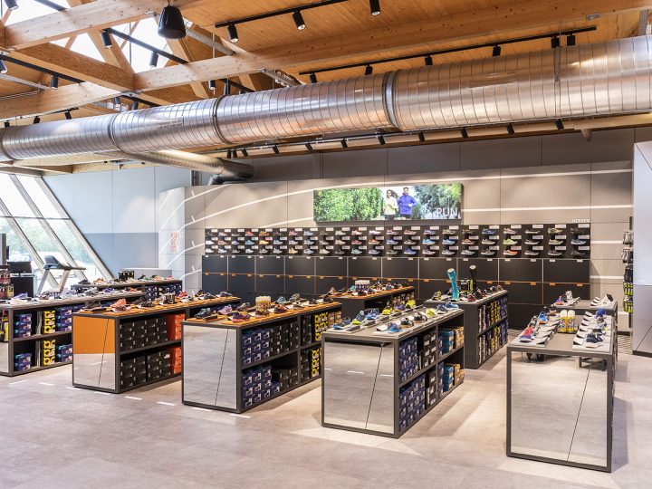 Un moderno negozio di scarpe con soffitti in legno, tubature a vista e grandi finestre. Espositori di scarpe con vari stili di scarpe sono sparsi per tutto lo spazio. Uno schermo digitale sulla parete di fondo mostra un'immagine di persone all'aperto.