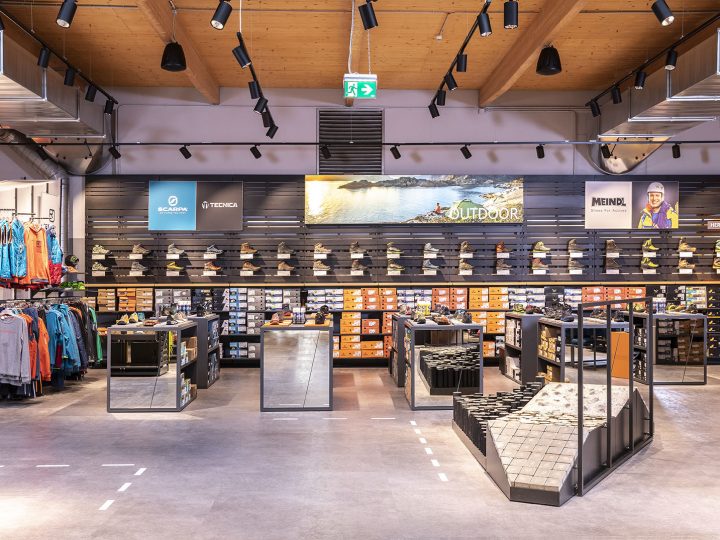 Un moderno negozio di scarpe con una selezione di scarpe da outdoor sugli scaffali. I marchi sono visibili su un display a parete. Il negozio presenta un'illuminazione elegante, accessori a specchio e un'area per provare le scarpe su una rampa strutturata. L'abbigliamento si trova sul lato sinistro.