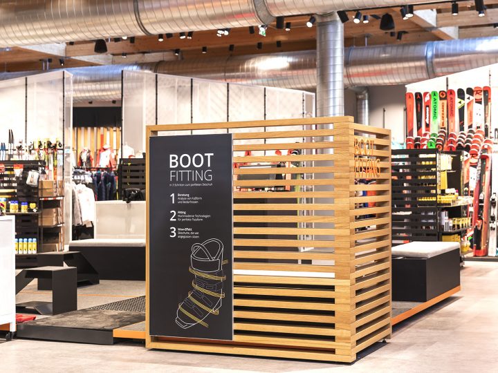 Un'area di prova scarponi in un negozio di sci con un elegante rivestimento in legno e un cartello nero. Sullo sfondo, varie attrezzature da sci, tra cui sci e bastoncini, sono ordinatamente disposte su scaffali.