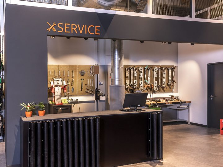 Negozio di ski service con bancone frontale, attrezzi montati a parete e una postazione di lavoro sullo sfondo. A sinistra, l'attrezzatura da sci è appesa a dei supporti. Sopra il bancone campeggia la parola "SERVICE".
