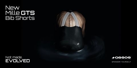 Una persona è vista da dietro con indosso dei pantaloncini da ciclista, nell'angolo in alto a sinistra c'è il testo "New Mille GTS Bib Shorts". L'immagine ha uno sfondo scuro che mette in risalto le bretelle riflettenti e il materiale dei pantaloncini.