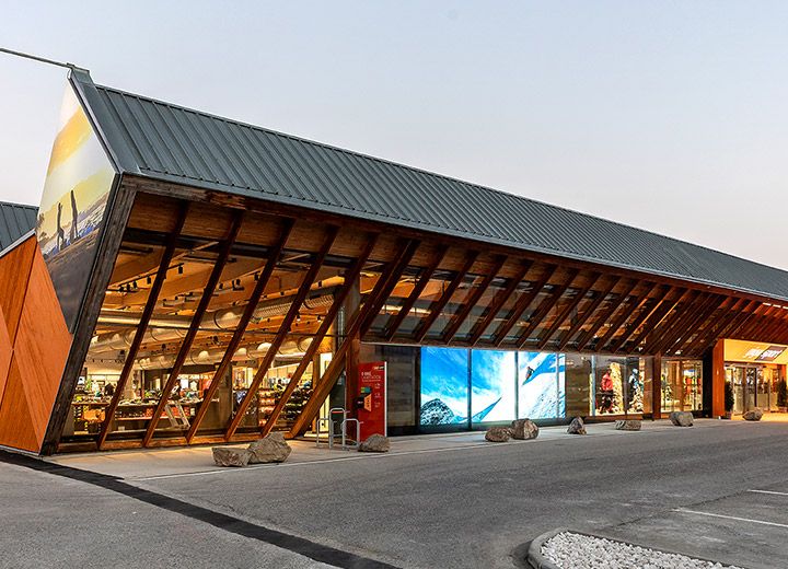 Un edificio moderno con un'esclusiva struttura a tetto spiovente, ampie vetrate ed elementi in legno, sullo sfondo di un cielo limpido. Nell'angolo in alto a destra si trova un cartello con la scritta "Vincitore del German Design Award 2020". L'esterno è illuminato con luce calda.