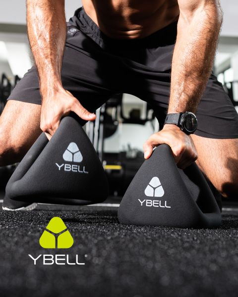 Una persona in abbigliamento da allenamento tiene due pesi YBELL neri sul pavimento della palestra. Indossa un orologio e dei pantaloncini. Il logo YBELL è raffigurato sui pesi e nell'angolo inferiore sinistro.