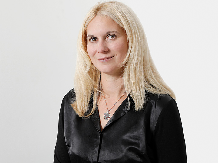 Una persona con lunghi capelli biondi, che indossa una blusa nera e una collana con un ciondolo rotondo, è in piedi davanti a uno sfondo bianco semplice e sorride leggermente.