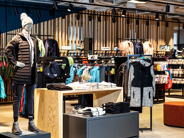 Interno di un negozio di abbigliamento con un manichino che indossa una giacca nera, un cappello bianco e stivali. Diverse giacche colorate e abbigliamento sportivo sono esposti su stand e scaffali, con accenti in legno e illuminazione moderna sullo sfondo.