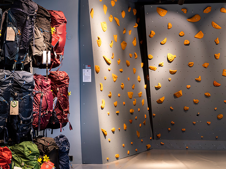Parete da arrampicata indoor con diverse prese arancioni su sfondo grigio. A sinistra, su scaffali, sono esposti diversi zaini di vari colori. L'illuminazione è intensa e il pavimento è lucido.