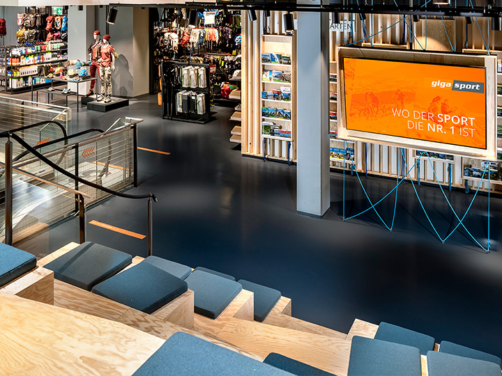 Interno di un negozio di articoli sportivi con sedute in legno con cuscini, un grande schermo con le scritte "Gigasport" e "Dove lo sport è il numero 1", scale mobili e articoli sportivi esposti. L'ambiente è moderno e ben illuminato.