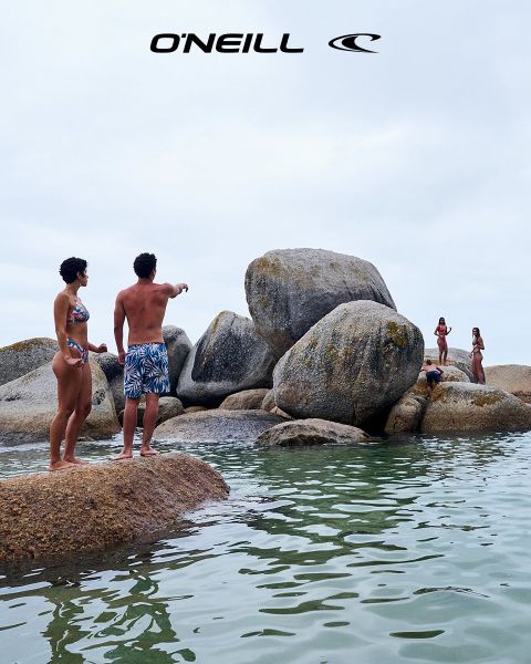 Una donna con i capelli lunghi si trova in un paesaggio desertico sotto un cielo blu con nuvole sparse. Indossa una canotta nera O'Neill e si sporge leggermente in avanti. Sul lato destro dell'immagine si vede il logo O'Neill.