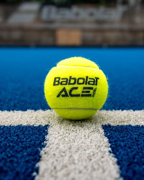 Una pallina da tennis Babolat gialla si trova su un campo da tennis blu, centrata su una linea bianca. Lo sfondo è sfocato e mette a fuoco il logo del marchio e la texture della pallina.