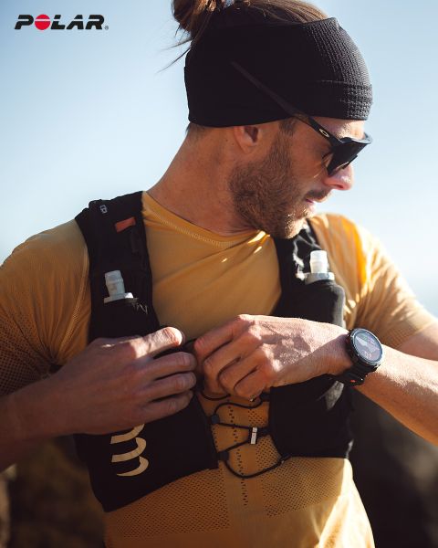 Una persona con una fascia nera e occhiali da sole si sistema il suo gilet da idratazione nero. Indossa una camicia gialla e uno smartwatch al polso. Il logo Polar è visibile in alto a sinistra. La luce del sole illumina la scena.