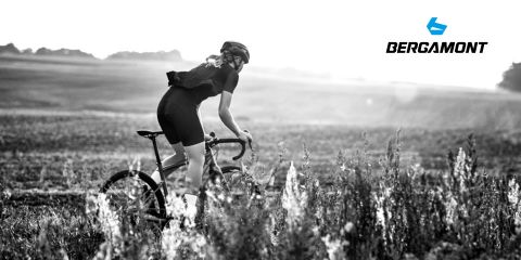 Un ciclista con casco sta attraversando un prato con una bici da corsa, sullo sfondo si vede un paesaggio collinare. L'immagine è in bianco e nero e nell'angolo in alto a destra si vede il logo Bergamont.