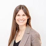 Donna sorridente con lunghi capelli castani, indossa un blazer a quadri e un top nero, in piedi davanti a uno sfondo bianco semplice.