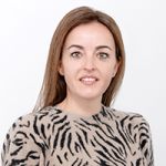 Una donna con i capelli castani sorride alla fotocamera. Indossa un maglione beige con un motivo a strisce zebrate nere. Lo sfondo è semplicemente bianco.