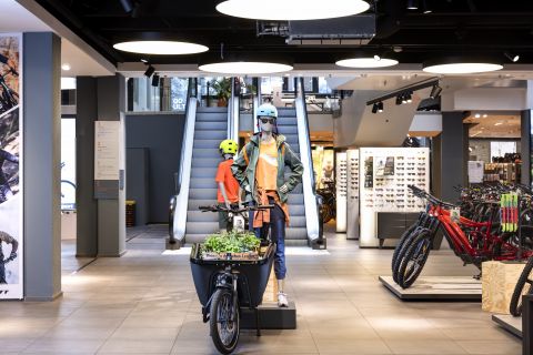 Eine Schaufensterpuppe in Fahrradkleidung steht mit einem Fahrrad und trägt einen Korb mit Gemüse in einem modernen Geschäft. Im Hintergrund sind eine Rolltreppe und Fahrradzubehör zu sehen. Das Geschäft ist hell beleuchtet und großzügig gestaltet.