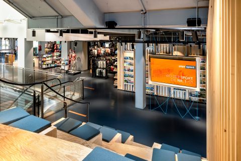 Innenansicht eines modernen Sportgeschäfts mit Sitzbereich mit Holzstufen und blauen Kissen. Auf einem großen orangefarbenen Bildschirm steht „Wo der Sport die Nr. 1 ist“. Das Geschäft ist gut beleuchtet und bietet verschiedene Sportgeräte und -bekleidung.