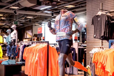 Eine Schaufensterpuppe in Sportkleidung, mit geblümter Jacke, schwarzen Shorts und orangefarbenen Kniestrümpfen, posiert in Laufposition in einem Sportbekleidungsgeschäft. Im Vordergrund sind leuchtend orangefarbene Shirts auf einem Ständer ausgestellt.