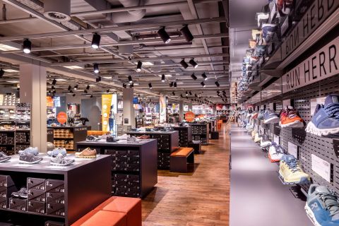 Ein geräumiges Schuhgeschäft mit übersichtlich angeordneten Reihen von Turnschuhen auf Regalen. Es gibt schwarze Ständer mit Schuhkartons und orangefarbene Sitzbänke für die Kunden. Das Geschäft ist gut beleuchtet und hat eine moderne Industriedecke.